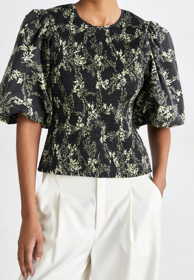 Femme portant un chemisier noir à motif floral beige avec des manches courtes bouffantes, associé à un pantalon blanc taille haute, mains dans les poches.