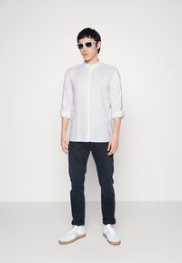 Weißes Leinenhemd mit Stehkragen, hochgekrempelten Ärmeln, kombiniert mit dunklen Jeans und weißen Sneakern. Das Model trägt weiße Sonnenbrillen.