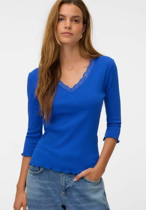 Femme aux longs cheveux bruns porte un haut bleu côtelé à col en V avec bordure en dentelle et manches trois-quarts, associé à un jean bleu clair.
