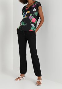 Blusa negra con estampado floral tropical, de mangas cortas y detalles de lazo; combinada con pantalones negros y sandalias de tacón cuadrado blancas.