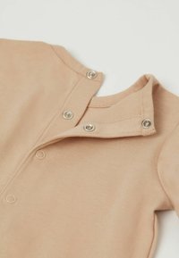 Beige Baumwoll-Overall mit Druckknöpfen am Kragen und Oberteil, gefertigt aus einem weichen, strukturierten Stoff und mit Rundhalsausschnitt.