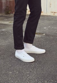 Nogi w czarnych podwiniętych spodniach, białych skarpetkach i białych sneakersach Vans stojące na asfalcie obok zniszczonej ściany.