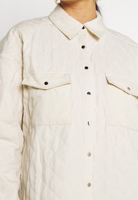 Camisa chaqueta acolchada de color beige claro con un cuello clásico, botones a presión y dos bolsillos en el pecho con tapas. Detalle de patrón de diamantes texturizado.