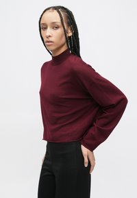 Top burgundy cu mâneci lungi și guler înalt, realizat dintr-un material moale, cu croială lejeră și umeri căzuți, asortat cu pantaloni negri.