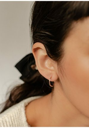 Gros plan sur l'oreille d'une femme aux cheveux foncés attachés en arrière par un chouchou noir, portant une petite boucle d'oreille en or.