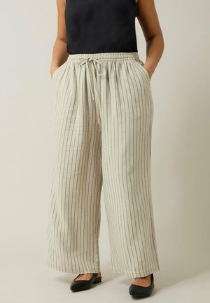 Person iført løse beige bukser med lodrette sorte striber, snøre omkring taljen, sort ærmeløs top og sorte sko med lav hæl.