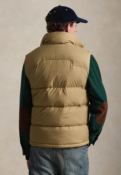 Hombre que lleva un chaleco acolchado color beige, camisa verde de manga larga con parches marrones en los codos, vaqueros azules y gorra de pana azul marino, de espaldas.