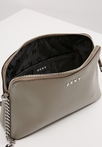 Borsa a mano sintetica grigia con esterno testurizzato, chiusura con zip e dettagli in metallo argentato. L'interno presenta una fodera nera con motivo del logo DKNY.