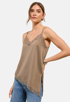 Jonge vrouw die een taupekleurige mouwloze zijden top met kanten rand en een blauwe spijkerbroek draagt, tegen een effen lichte achtergrond.