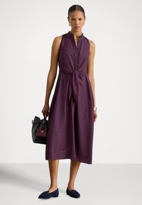 Ermeløs kjole til knelengde i lilla med geometrisk mønster, med et knytebånd i midjen. Kombinert med marineblå loafers og en svart håndveske.