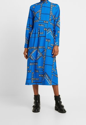 Blaues Midikleid mit geometrischen und floralen Mustern, langen Ärmeln, Kragen, getragen mit schwarzen Stiefeletten mit Schnalle.