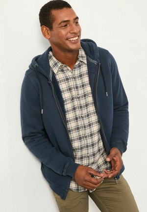 Marineblauer Reißverschluss-Hoodie mit frontalen Taschen über einem karierter Kurzarmhemd. Verfügt über strukturierten Stoff und eine lässige Passform. Olivgrüne Hose sichtbar.