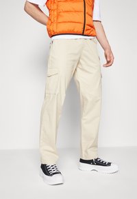 Pantalon cargo beige avec poches latérales, associé à un t-shirt blanc et un gilet matelassé orange, porté avec des baskets noires et blanches.