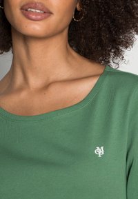 Zelené, pruhované tričko s širokým výstřihem a bílým logem na hrudi. Materiál vypadá měkce a těsně přiléhavě.