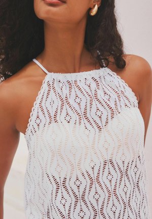 Débardeur blanc sans manches avec des motifs découpés complexes, des lignes ondulées et des bords festonnés. Les bretelles sont fines et délicates.