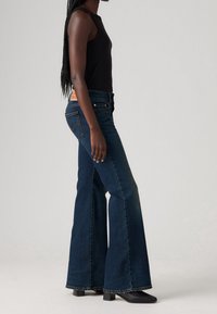 Femme portant un jean flare taille haute bleu foncé, un haut noir sans manches et des bottines noires à talons, debout de profil.