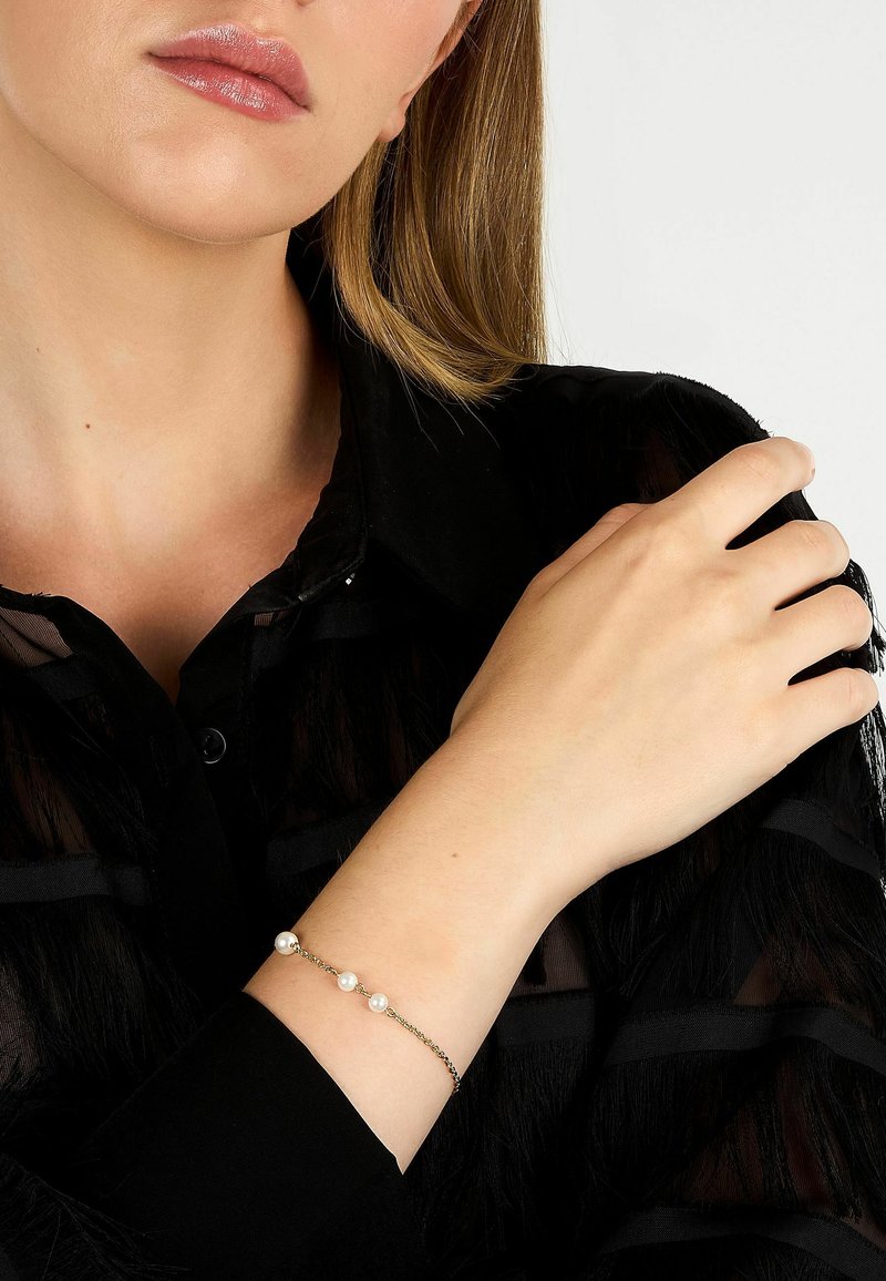 Silverfärgat kedjearmband med tre vita pärlor, bärs på en handled. Bakgrunden visar en svart, genomskinlig, strukturerad skjorta.