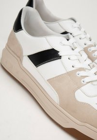 Bílé a béžové tenisky s černými akcenty. Mají hladkou kůži a semišové textury, design na šněrování a gumovou podrážku s gripem.