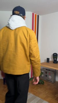 Veste bomber jaune avec doublure à capuche grise, associée à un pantalon noir. En arrière-plan, un mur rayé coloré et des meubles en bois.