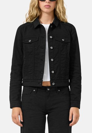 Frau trägt schwarze Jeansjacke mit silbernen Knöpfen, schwarze Jeans, weißes bauchfreies Top und schwarze Sonnenbrille vor schlichtem Hintergrund.