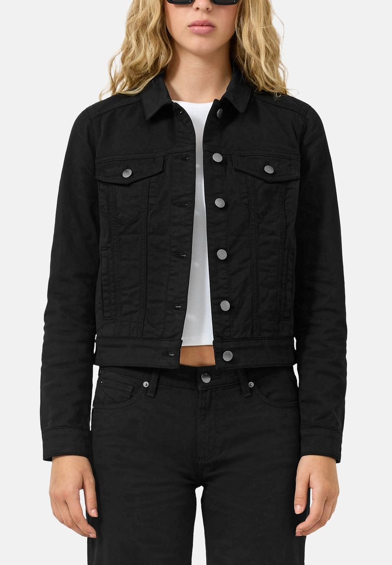 Vrouw draagt een zwart denimjack met zilveren knopen, zwarte spijkerbroek, wit cropped topje en zwarte zonnebril tegen een effen achtergrond.