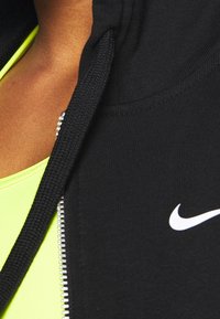 Gros plan d'une personne portant un sweat à capuche zippé noir Nike par-dessus un haut jaune vif.