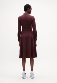 Rochie burgundă cu mâneci lungi, guler, detaliu cu brâu și fustă cloș, decorată cu nasturi pe manșete. Este purtată cu adidași albi.