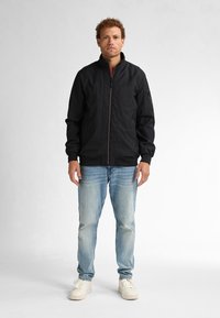 Giacca bomber nera con zip frontale, polsini a coste e tasca laterale. Indossata con jeans azzurri e sneakers bianche, in piedi davanti a uno sfondo neutro.