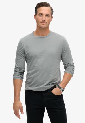 Camiseta gris de manga larga hecha de una tela suave, con un escote tipo crew, combinada con jeans negros y un reloj en la muñeca.