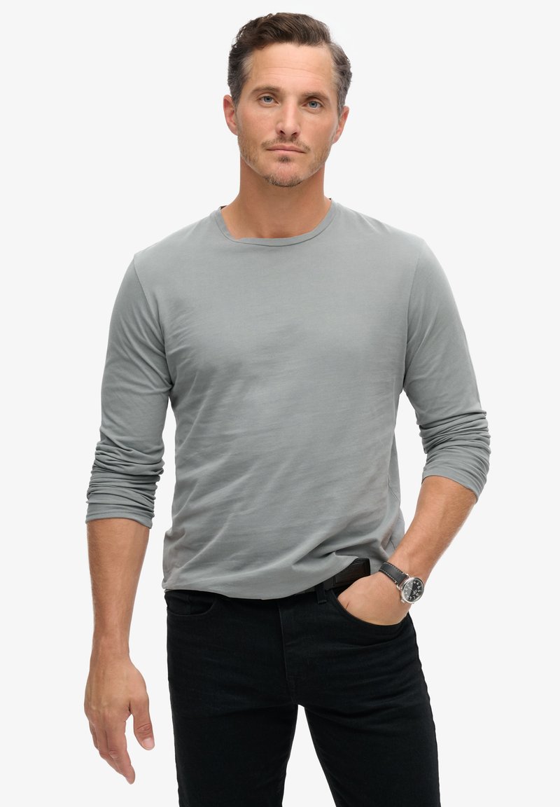 Grijze lange mouwen t-shirt gemaakt van zachte stof, met een ronde hals, gecombineerd met zwarte jeans en een horloge om de pols.