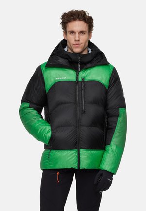 Man draagt een zwart-groene donsjas met kap, staat met één hand in de zak en de andere in een zwarte handschoen.