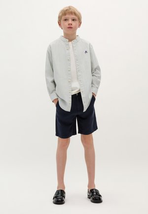 Camicia a righe blu chiaro e bianco, abbinata a pantaloni corti blu navy e scarpe nere. La camicia presenta un colletto alla cinese e un logo sul petto.