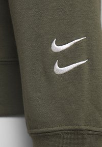 Zelená látka mikiny s texturovaným vzhledem; má dvě bílé vyšívané loga Nike na rukávu.