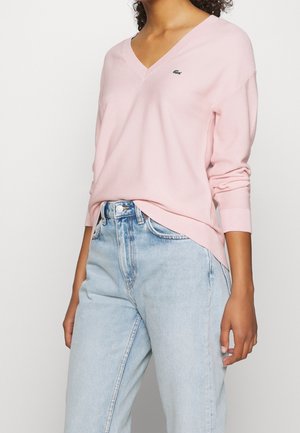 Femme portant un pull rose clair col V avec un petit logo vert et un jean bleu clair taille haute sur un fond uni.