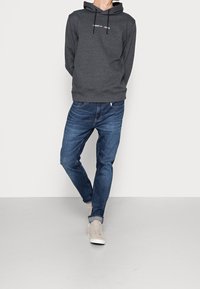 Personne portant un sweat à capuche Tommy Jeans gris foncé, un jean bleu retroussé et des sneakers beige clair, se tenant contre un mur blanc.