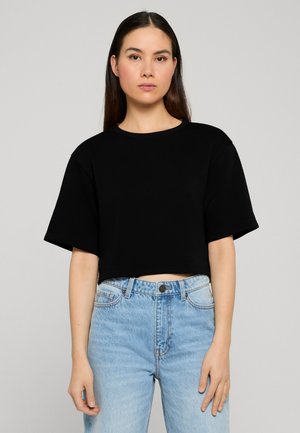 Ung kvinde med glat mørkt hår iført en sort cropped t-shirt og lyseblå højtaljede jeans mod en ensfarvet baggrund.
