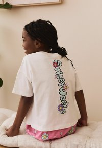 T-shirt en coton blanc avec des graphiques floraux colorés et le texte "SMILEY WORLD" dans le dos. Porté avec un short rose à motifs floraux.