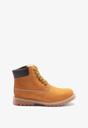Botte chelsea en daim beige avec un col rembourré noir, des œillets dorés et des coutures beiges. Elle présente une épaisse semelle en caoutchouc avec des rainures de traction.