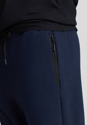 Granatowe joggery wykonane z miękkiego materiału, z boczną kieszenią na zamek w czarnym kolorze, elastycznym pasem z sznurkiem oraz prostym krojem.