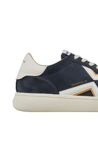 Sneaker in suede blu navy con accenti bianchi e suola in gomma beige. Presenta il marchio dorato "SMITH X" e un design geometrico sul lato.