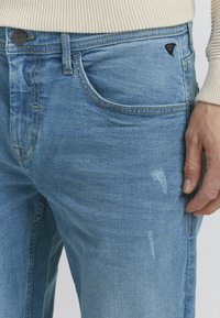 Jeans de mezclilla azul claro con una textura desgastada, que cuentan con un bolsillo frontal y un pequeño logo negro en la cinturilla. Incluye costuras visibles.