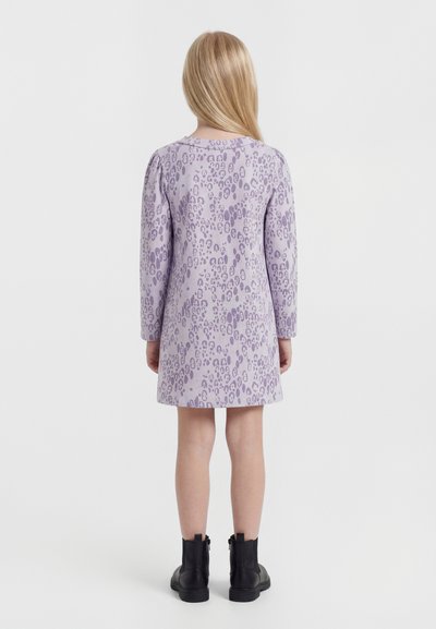 Παιδί με μακριά ξανθά μαλλιά που φοράει μωβ φόρεμα με λεοπάρ σχέδιο και μαύρα μποτάκια μέχρι τον αστράγαλο, στέκεται γυρισμένο με την πλάτη σε λευκό φόντο.