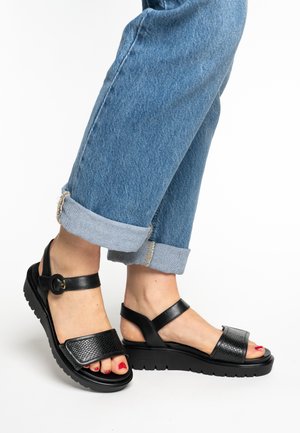 Black leather sandals with textured strap, adjustable ankle buckle, and chunky sole. Worn with rolled-up blue denim jeans.