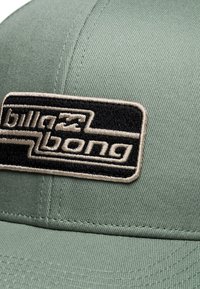 Cappello verde con superficie testurizzata e visiera piatta. Presenta un distintivo logo ricamato Billabong in nero e beige sulla parte anteriore.