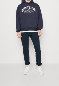 Námořnická mikina s kapucí s bílým nápisem, přední kapsou a žebrovanými manžetami, v kombinaci s tmavými kalhotami a bílými teniskami. Jednoduchý design.