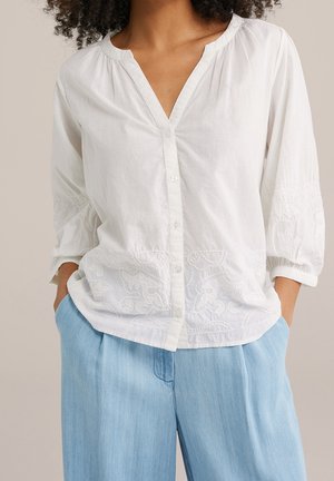 Kobieta w białej haftowanej koszuli zapinanej na guziki z rękawami 3/4 i luźnych, jasnoniebieskich spodniach, z rękami w kieszeniach.