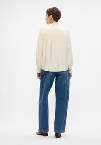 Blusa a maniche lunghe color crema con dettaglio arricciato sulla schiena, accenti di pizzo sui polsini, abbinata a jeans blu larghi e stivali con tacco marroni.