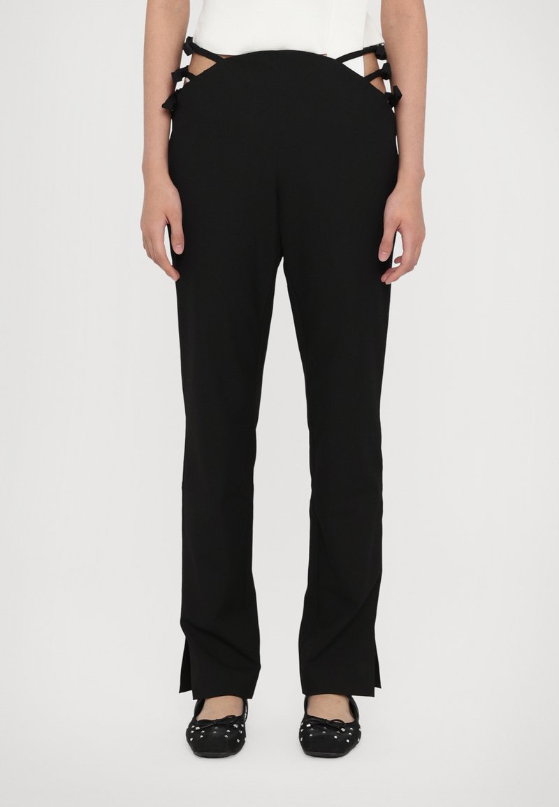 Zwarte broek met een gladde textuur, heeft uitsnijdingen aan de zijkanten die zijn vastgebonden met Stoffen linten, en splitten aan de zoom. Gecombineerd met zwarte platte schoenen.