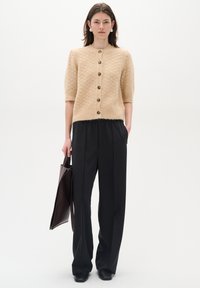 Beige pletený cardigan s krátkymi rukávmi, textúrovaná tkanina, päť hnedých gombíkov, v kombinácii s vysokým čiernym pásom a hnedou taškou.