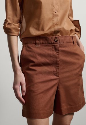 Persoon draagt een opgerold bruinen lange-mouwen overhemd, ingestopt in een roestkleurige short met hoge taille, voorzien van een knoop en zijzakken.